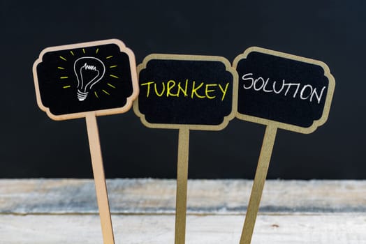 Concept message TURNKEY SOLUTION and light bulb as symbol for idea written with chalk on wooden mini blackboard labels, defocused chalkboard and wood table in background