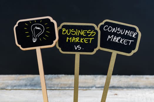 Concept message BUSINESS MARKET versus CONSUMER MARKET and light bulb as symbol for idea written with chalk on wooden mini blackboard labels, defocused chalkboard and wood table in background