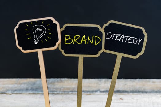 Concept message BRAND STRATEGY and light bulb as symbol for idea written with chalk on wooden mini blackboard labels, defocused chalkboard and wood table in background