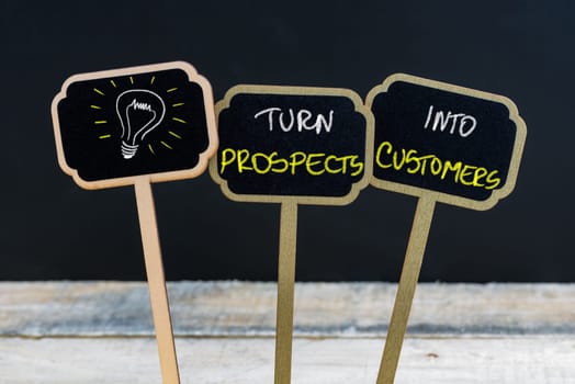 Concept message TURN PROSPECTS INTO CUSTOMERS and light bulb as symbol for idea written with chalk on wooden mini blackboard labels, defocused chalkboard and wood table in background