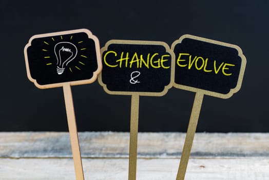 Concept message CHANGE AND EVOLVE and light bulb as symbol for idea written with chalk on wooden mini blackboard labels, defocused chalkboard and wood table in background