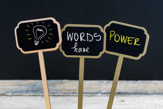 Concept message WORDS HAVE POWER and light bulb as symbol for idea written with chalk on wooden mini blackboard labels, defocused chalkboard and wood table in background