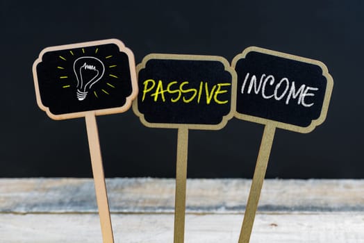 Concept message PASSIVE INCOME and light bulb as symbol for idea written with chalk on wooden mini blackboard labels, defocused chalkboard and wood table in background