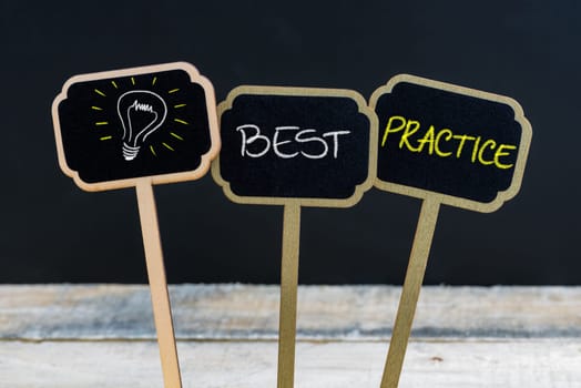 Concept message BEST PRACTICE and light bulb as symbol for idea written with chalk on wooden mini blackboard labels, defocused chalkboard and wood table in background