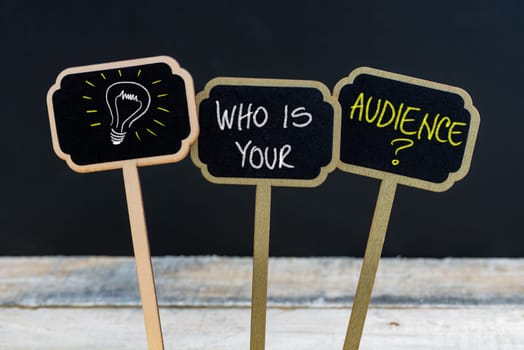 Concept message WHO IS YOUR AUDIENCE ? and light bulb as symbol for idea written with chalk on wooden mini blackboard labels, defocused chalkboard and wood table in background