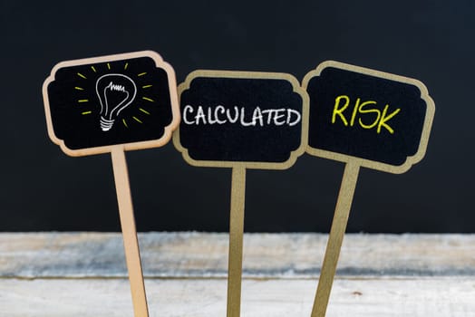 Concept message CALCULATED RISK and light bulb as symbol for idea written with chalk on wooden mini blackboard labels, defocused chalkboard and wood table in background