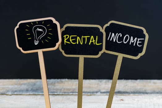 Concept message RENTAL INCOME and light bulb as symbol for idea written with chalk on wooden mini blackboard labels, defocused chalkboard and wood table in background