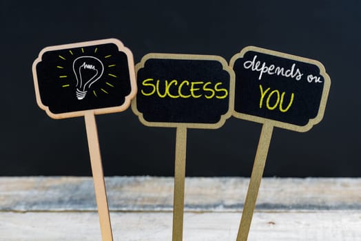 Concept message SUCCESS DEPENDS ON YOU and light bulb as symbol for idea written with chalk on wooden mini blackboard labels, defocused chalkboard and wood table in background