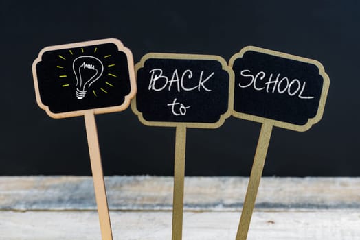 Concept message BACK TO SCHOOL and light bulb as symbol for idea written with chalk on wooden mini blackboard labels, defocused chalkboard and wood table in background