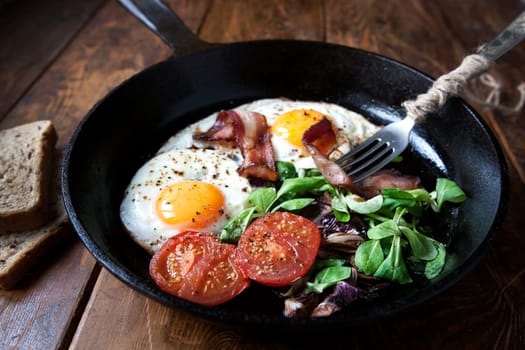 Breakfast set. Pan of fried eggs with bacon, fresh tomato, sage and bread on dark serving board over black wooden background, top view, copy space