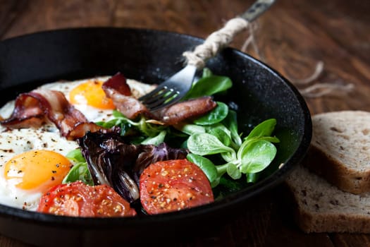 Breakfast set. Pan of fried eggs with bacon, fresh tomato, sage and bread on dark serving board over black wooden background, top view, copy space