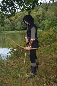 Young medieval archer with black hood stands at the lake with a tense bow and arrow
