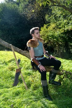 Young medieval dressed man,sitting on branch,hands on legs and looking to the side,bow and arrow stands next to it