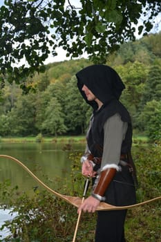 Young medieval archer with black hood stands at the lake with a tense bow and arrow