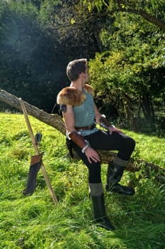Young man dressed in medieval clothes,sits on branch,hands on legs and looks backwards,bow and arrow stands next to it