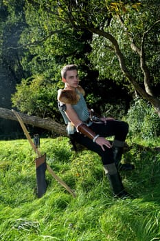Young medieval dressed man,sitting on branch,hands on legs and looking forward,bow and arrow stands next to it