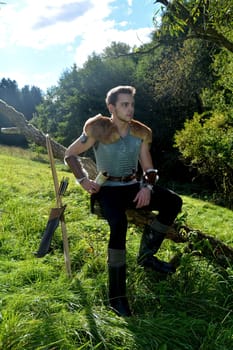 Young medieval archer with chain mail sits on branch in nature in the sunlight,bow and arrow stands next to it