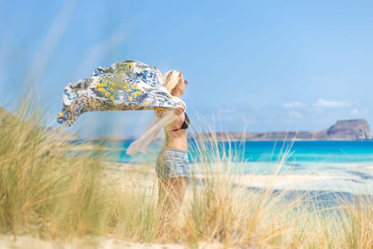 Relaxed woman, arms raised, holding colorful scarf, enjoying sun, freedom and life at beautiful beach. Young lady feeling free, relaxed and happy. Concept of vacations, freedom, joy and well being.