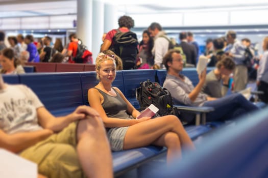Casual blond young woman waiting to board a plane at airport departure gates, holding flight ticket and passport in her hands.