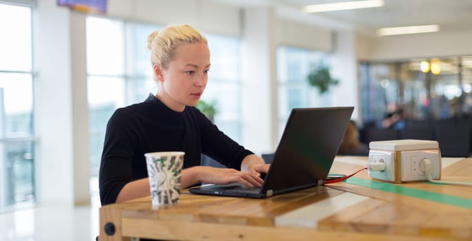 Female freelancer remotely working on the go using multiple devices on a desk at airport coffee shop while traveling. Contemporary entrepreneurial lifestyle.