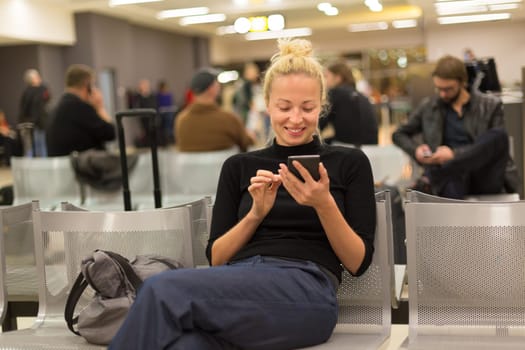 Casual blond young woman using her mobile phone while waiting to board a plane at airport departure gates.