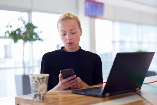 Female freelancer remotely working on the go using multiple devices on a desk at airport coffee shop while traveling. Contemporary entrepreneurial lifestyle.