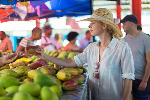 Female traveler wearing elegant colonial style white tunic and hat buying fresh tropical fruit on traditional Victoria food market on Seychelles islands.