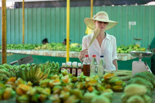 Female traveler wearing elegant colonial style white tunic and hat buying fresh tropical fruit on traditional Victoria food market on Seychelles islands.