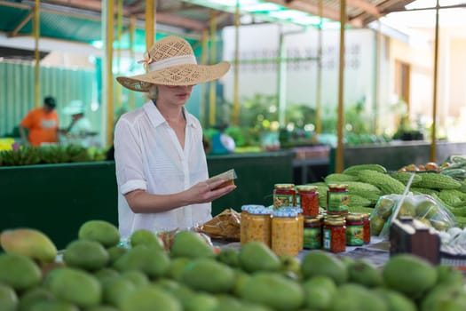 Female traveler wearing elegant colonial style white tunic and hat buying fresh tropical fruit on traditional Victoria food market on Seychelles islands.