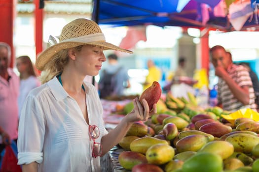 Female traveler wearing elegant colonial style white tunic and hat buying fresh tropical fruit on traditional Victoria food market on Seychelles islands.