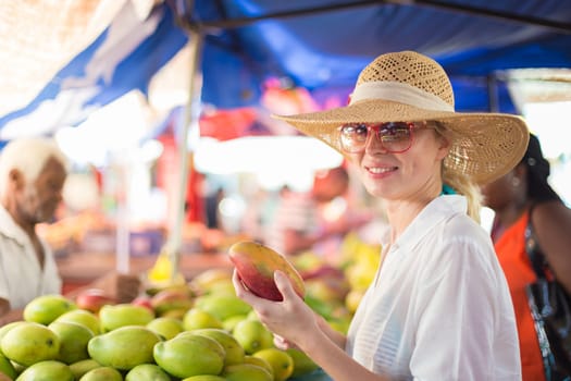 Female traveler wearing elegant colonial style white tunic and hat buying fresh tropical fruit on traditional Victoria food market on Seychelles islands.