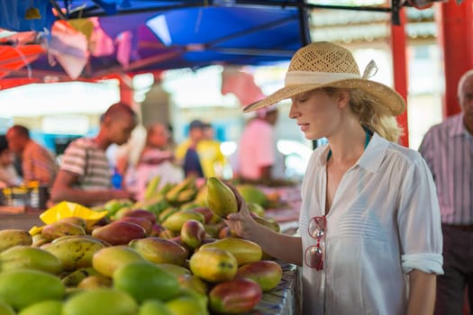 Female traveler wearing elegant colonial style white tunic and hat buying fresh tropical fruit on traditional Victoria food market on Seychelles islands.