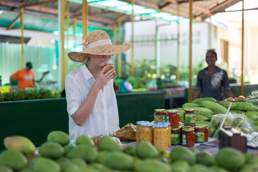 Female traveler wearing elegant colonial style white tunic and hat buying fresh tropical fruit on traditional Victoria food market on Seychelles islands.
