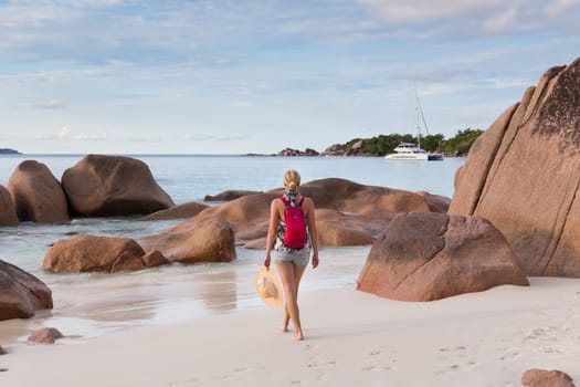 Woman wearing smal backpack, scarf, jeans shorts and beach hat, enjoying view of Anse Lazio beach on Praslin Island, Seychelles. Summer vacations on picture perfect tropical beach concept.
