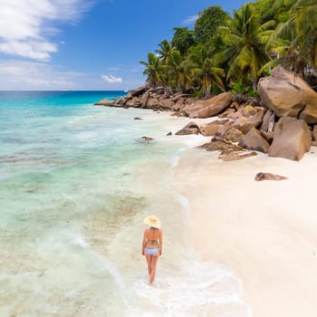 Woman wearing retro striped bikini and beach hat, enjoying amazing view on Anse Patates beach on La Digue Island, Seychelles. Summer vacations on picture perfect tropical beach concept.
