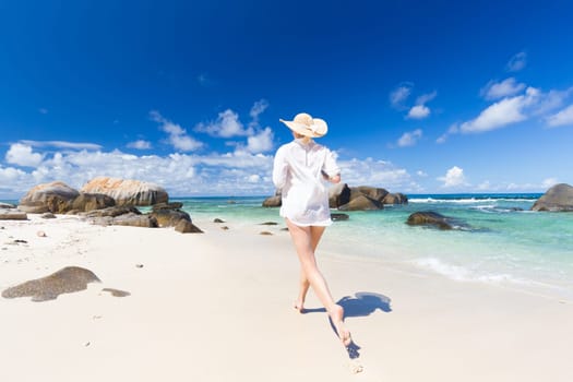 Active woman wearing white loose tunic over bikini and beach hat, enjoying amazing white sandy beach on Mahe Island, Seychelles. Summer vacations on picture perfect tropical beach concept.