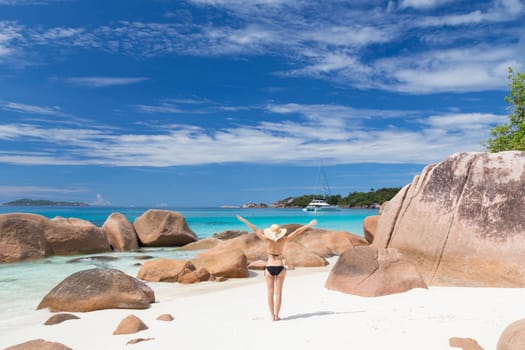 Woman arms rised, wearing black bikini and beach hat, enjoying amazing view on Anse Lazio beach on Praslin Island, Seychelles. Summer vacations on picture perfect tropical beach concept.
