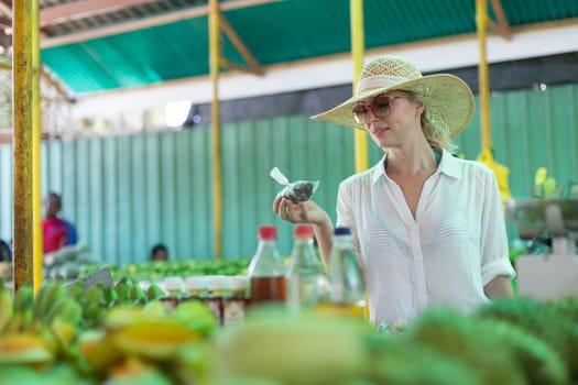 Female traveler wearing elegant colonial style white tunic and hat buying fresh tropical fruit on traditional Victoria food market on Seychelles islands.