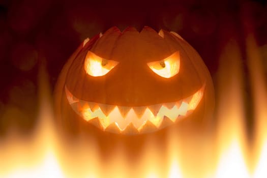 The big Helloween pumpkin has a face that laughs and is cheeky with shining eyes and a mouth with pointed teeth. Perspective from below in burning flames.