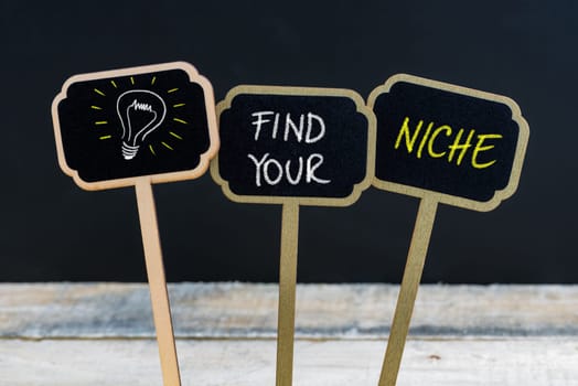 Concept message FIND YOUR NICHE and light bulb as symbol for idea written with chalk on wooden mini blackboard labels, defocused chalkboard and wood table in background