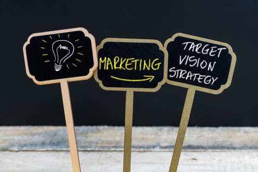 Concept message MARKETING - TARGET, VISION, STRATEGY and light bulb as symbol for idea written with chalk on wooden mini blackboard labels, defocused chalkboard and wood table in background