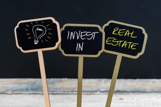 Concept message INVEST IN REAL ESTATE and light bulb as symbol for idea written with chalk on wooden mini blackboard labels, defocused chalkboard and wood table in background