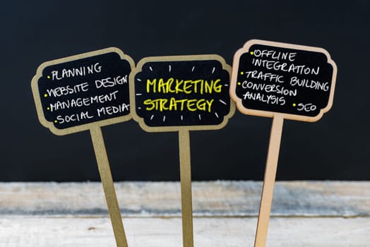 Concept message MARKETING STRATEGY written with chalk on wooden mini blackboard labels, defocused chalkboard and wood table in background