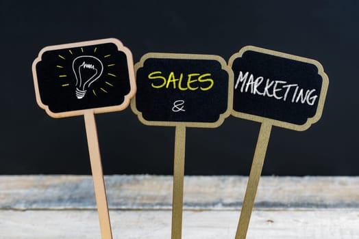 Concept message SALES AND MARKETING and light bulb as symbol for idea written with chalk on wooden mini blackboard labels, defocused chalkboard and wood table in background