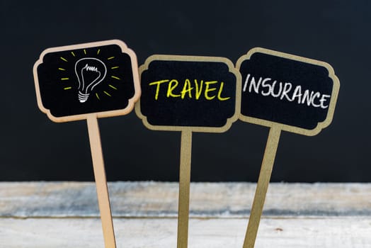 Concept message TRAVEL INSURANCE and light bulb as symbol for idea written with chalk on wooden mini blackboard labels, defocused chalkboard and wood table in background