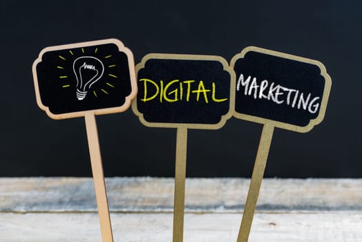 Concept message DIGITAL MARKETING and light bulb as symbol for idea written with chalk on wooden mini blackboard labels, defocused chalkboard and wood table in background