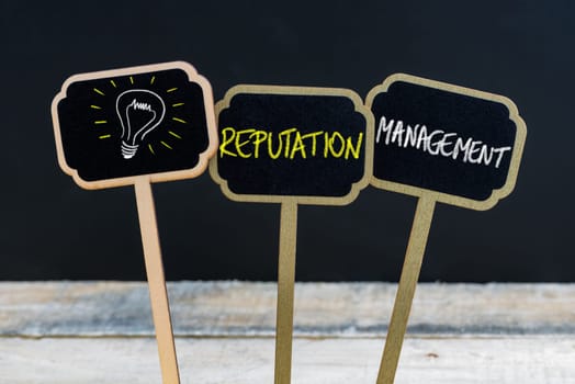 Concept message REPUTATION MANAGEMENT and light bulb as symbol for idea written with chalk on wooden mini blackboard labels, defocused chalkboard and wood table in background