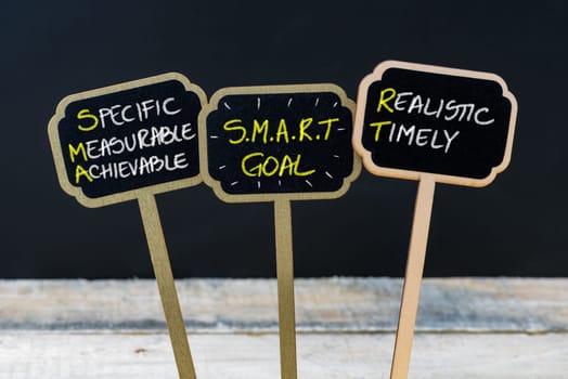 Concept message SMART GOAL written with chalk on wooden mini blackboard labels, defocused chalkboard and wood table in background