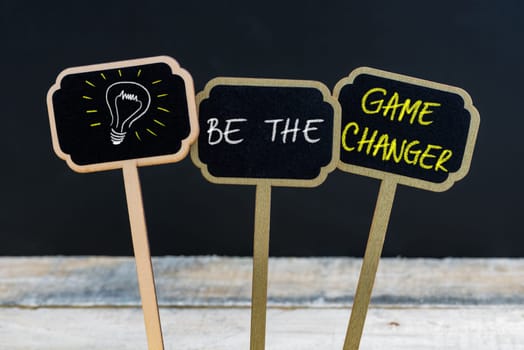 Concept message BE THE GAME CHANGER and light bulb as symbol for idea written with chalk on wooden mini blackboard labels, defocused chalkboard and wood table in background
