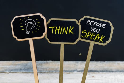 Concept message THINK BEFORE YOU SPEAK and light bulb as symbol for idea written with chalk on wooden mini blackboard labels, defocused chalkboard and wood table in background